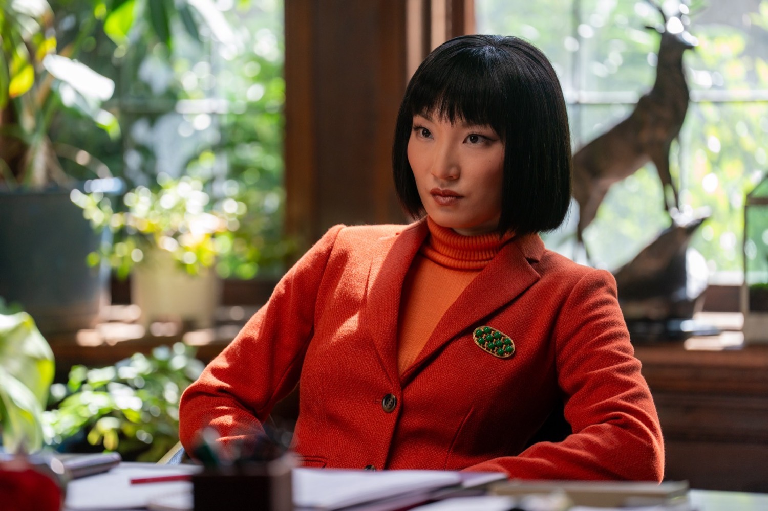 An actor in a red wool suit and orange turtleneck seated at a desk, His and Hers