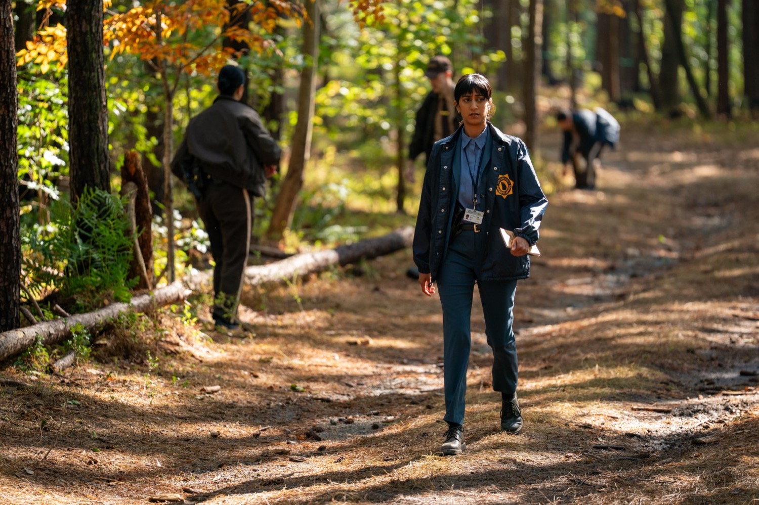 A detective in a navy field jacket walking a wooded crime scene, His and Hers